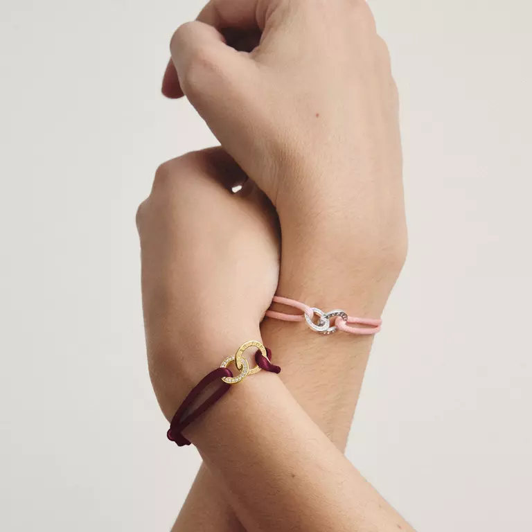 Three delicate charm bracelets displayed on a person's wrist, featuring a burgundy cord with a gold interlocking rings charm, a light pink cord with a silver interlocking rings charm, and a pink cord with a clear crystal charm.