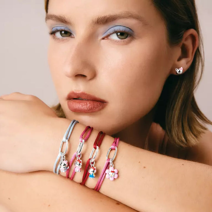 A close-up of a woman's face and crossed arms showing several colorful charm bracelets on her wrist. The bracelets feature silver-tone chain links and charms including a light blue cat, a red cat, a pink cat, and a small blue and white character charm.