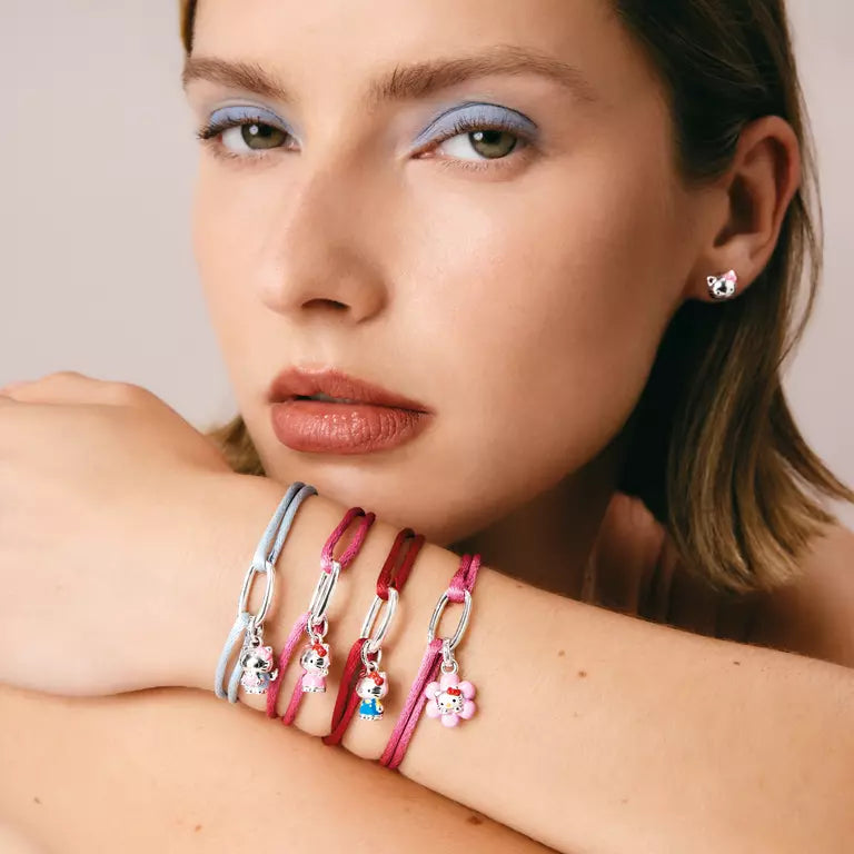 A close-up of a woman's face and crossed arms showing several colorful charm bracelets on her wrist. The bracelets feature silver-tone chain links and charms including a light blue cat, a red cat, a pink cat, and a small blue and white character charm.