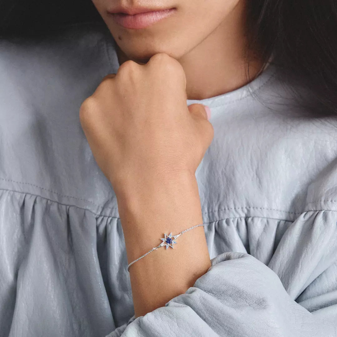 A close-up of a model's wrist wearing a delicate silver chain bracelet with a star-shaped centerpiece set with deep blue gemstones, paired with a light blue blouse.