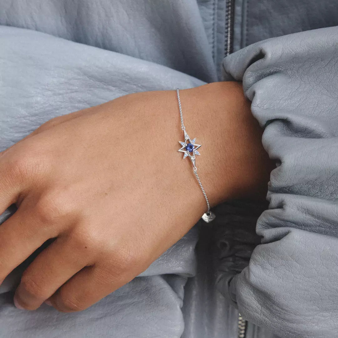 A close-up of a model's wrist wearing a delicate silver chain bracelet with a star-shaped centerpiece set with deep blue gemstones, paired with a light blue blouse.