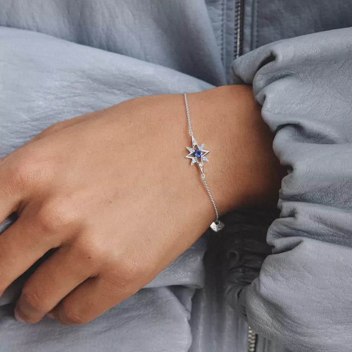 A close-up of a model's wrist wearing a delicate silver chain bracelet with a star-shaped centerpiece set with deep blue gemstones, paired with a light blue blouse.