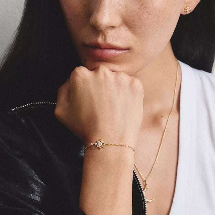 A close-up lifestyle shot of a model wearing a delicate gold chain bracelet with a small starburst design set with clear stones. The model is wearing a white top and a black leather jacket, with a gold necklace also visible.