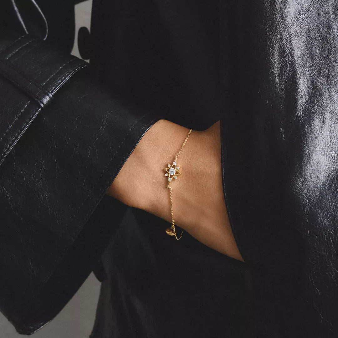 A delicate gold chain bracelet with a flower and crescent moon charm, worn on a person's wrist against a black leather jacket.