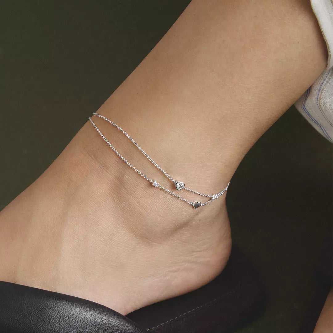 A close-up of a person's ankle wearing a delicate double-strand silver chain anklet with small star and heart charms.