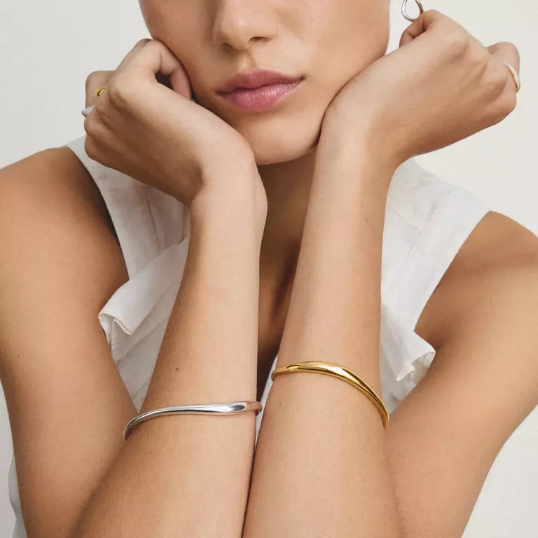 A close-up lifestyle shot of a model wearing two minimalist bangle bracelets on her arms, one in polished silver and one in polished gold, against a clean white background.