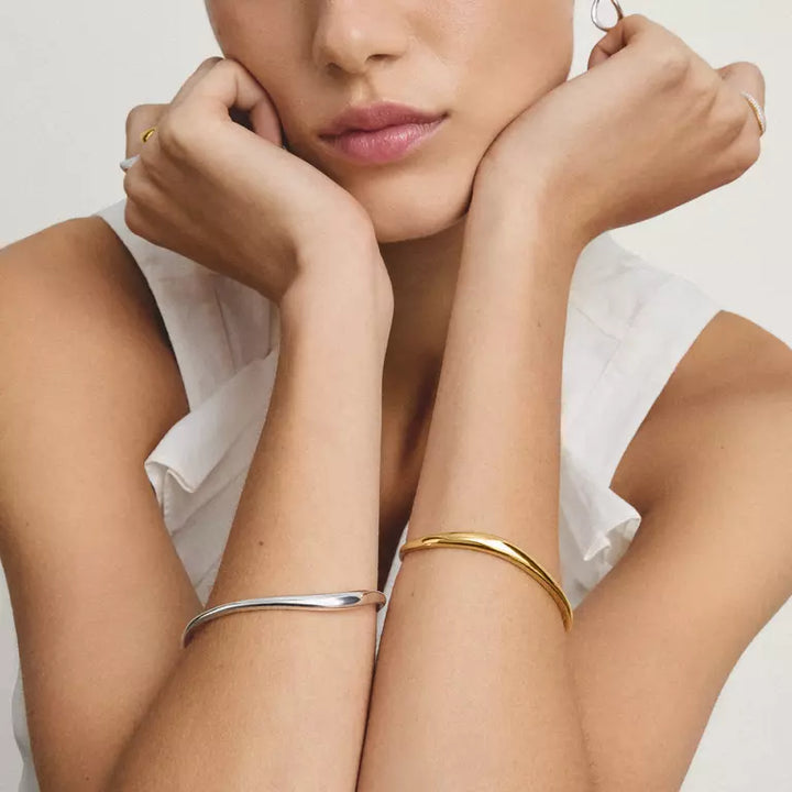 A close-up lifestyle shot of a model wearing two minimalist bangle bracelets on her arms, one in polished silver and one in polished gold, against a clean white background.