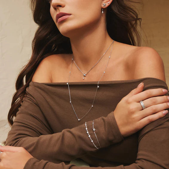 Woman wearing a brown off-shoulder top with silver jewelry including necklaces, bracelets, and rings.