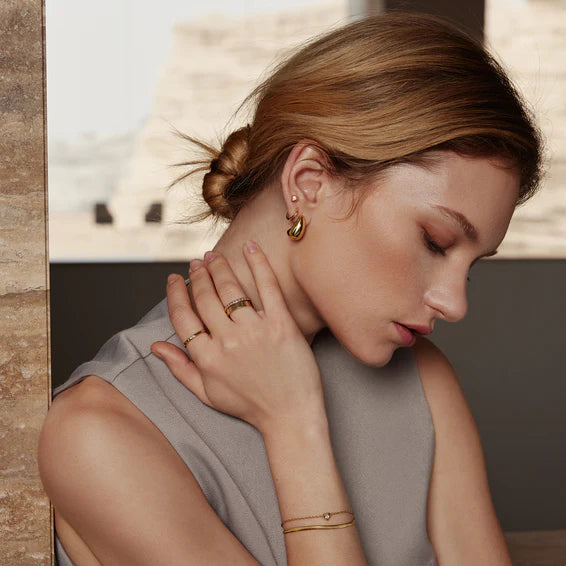 Woman wearing gold jewelry including earrings, rings, and a bracelet against a neutral background