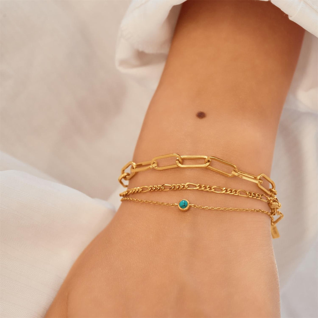 Gold chain bracelet with a turquoise bead on a wrist against a neutral background