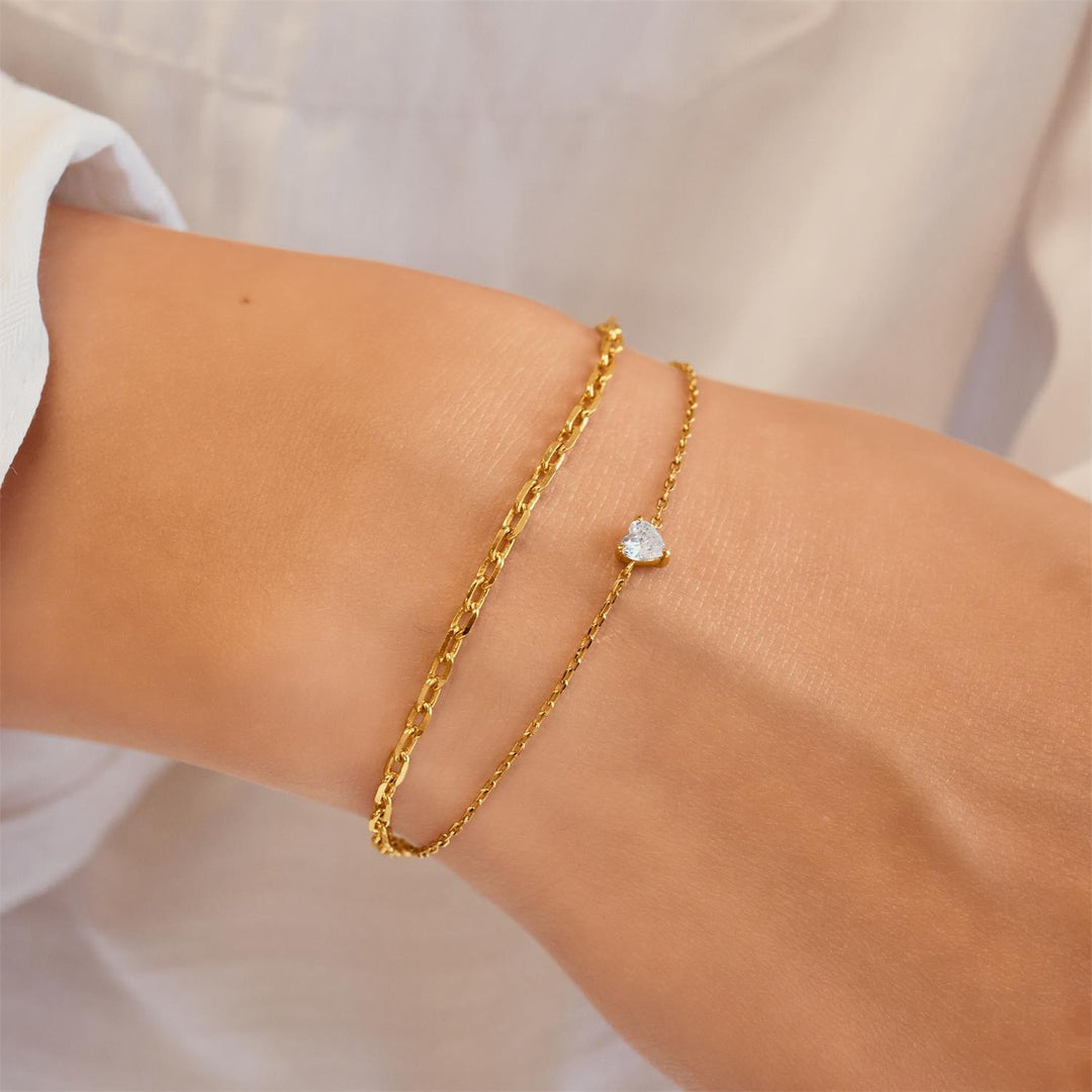 Gold bracelet with a heart-shaped charm on a wrist against a neutral background