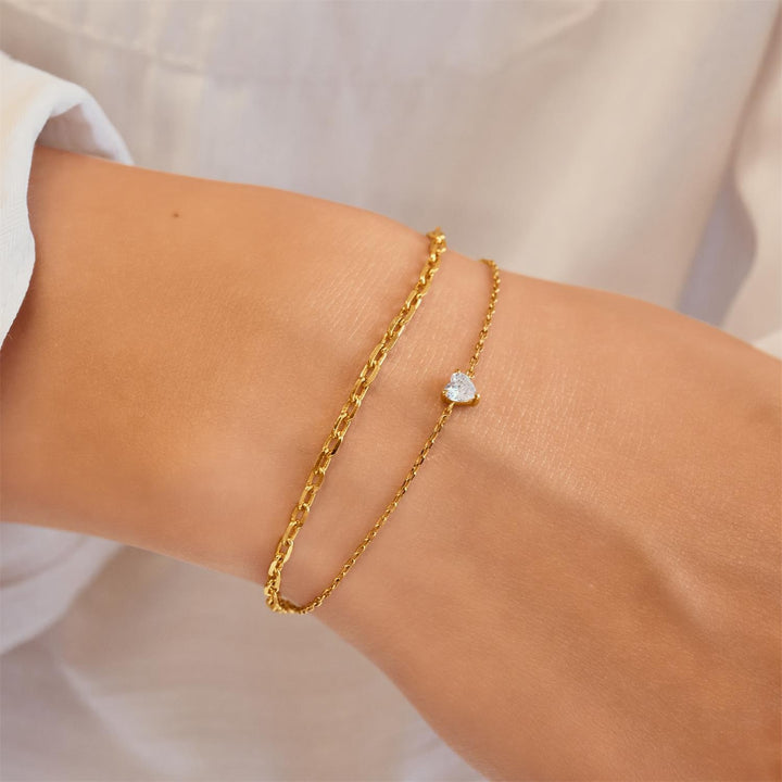 Gold bracelet with a heart-shaped charm on a wrist against a neutral background
