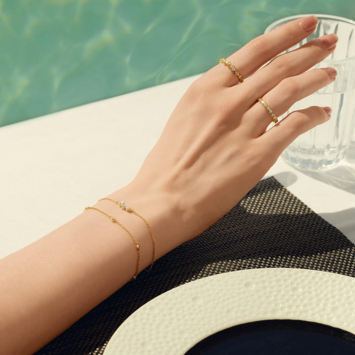 Hand wearing gold rings and bracelets with a blurred pool background