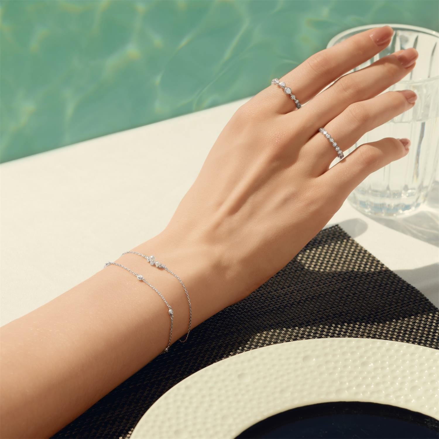 Hand wearing silver rings and bracelet with a blurred pool background