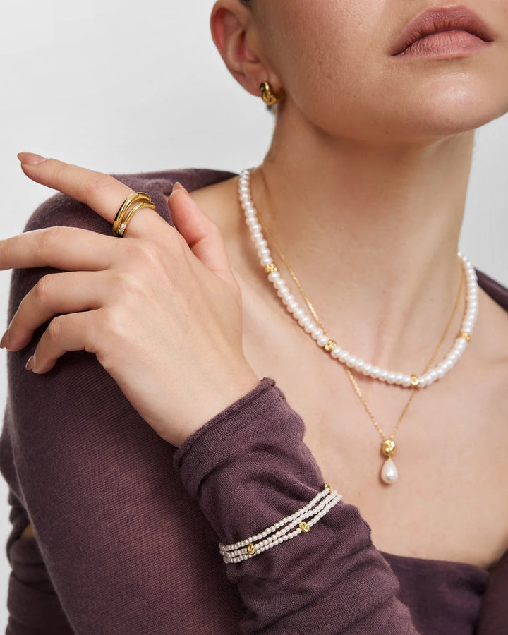 Close-up of a model wearing layered jewelry, including a double-strand pearl necklace with gold spacer beads, a gold teardrop pendant necklace, a gold ring set, and a pearl-and-gold bracelet on a mauve long-sleeve top.