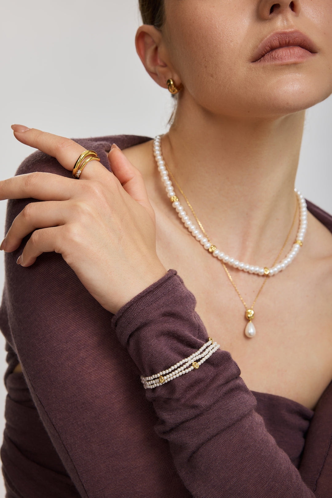 Close-up of a model wearing layered pearl and gold necklaces, a pearl bracelet, and gold rings, wearing a dark plum long-sleeve top against a light gray background.