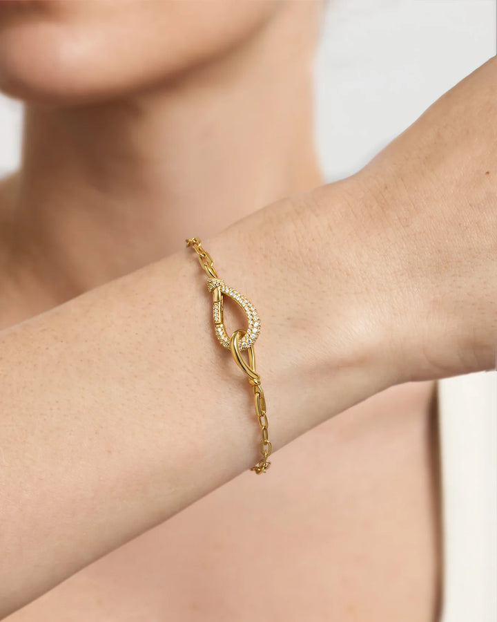 A close-up of a delicate gold chain bracelet with a central knot design and small clear stones, worn on a person's wrist.