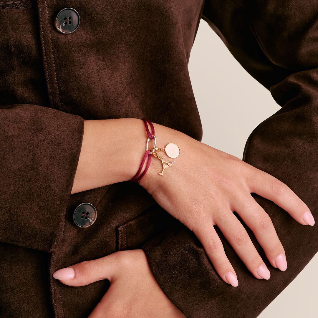 Close-up of a person wearing a gold bracelet with a letter 'A' charm on a neutral background.