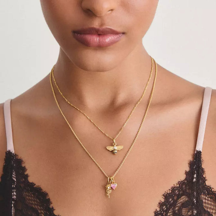 A close-up of a model wearing two layered gold necklaces. The shorter chain features a small bee pendant, while the longer chain has a rose pendant with a pink heart-shaped gemstone.