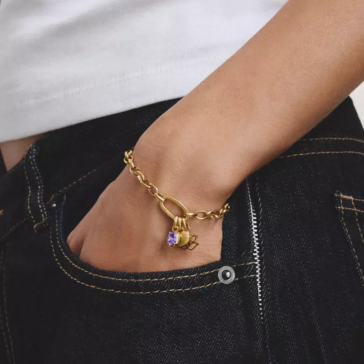 A gold chain bracelet with a small purple crystal charm and a tiny charm, worn on a person's wrist with a hand in a black denim pocket.