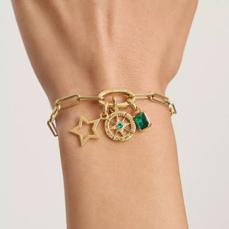 A gold chain bracelet with charms worn on a wrist, featuring a star charm, a compass wheel charm with a small green stone, and a rectangular green gem charm.