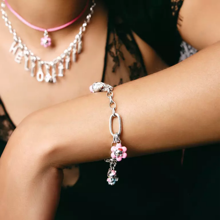 A silver chain bracelet with a snake charm and pink flower charms worn on a woman's wrist, with a silver letter necklace and pink choker in the background.