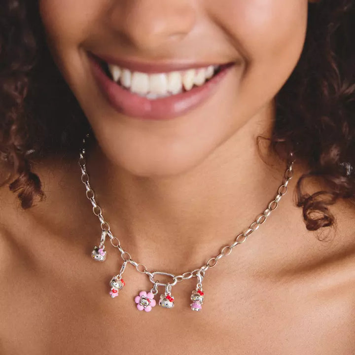 A close-up of a smiling woman wearing a silver chain necklace with dangling flower charms in pink, red, and white, with small rhinestone accents.