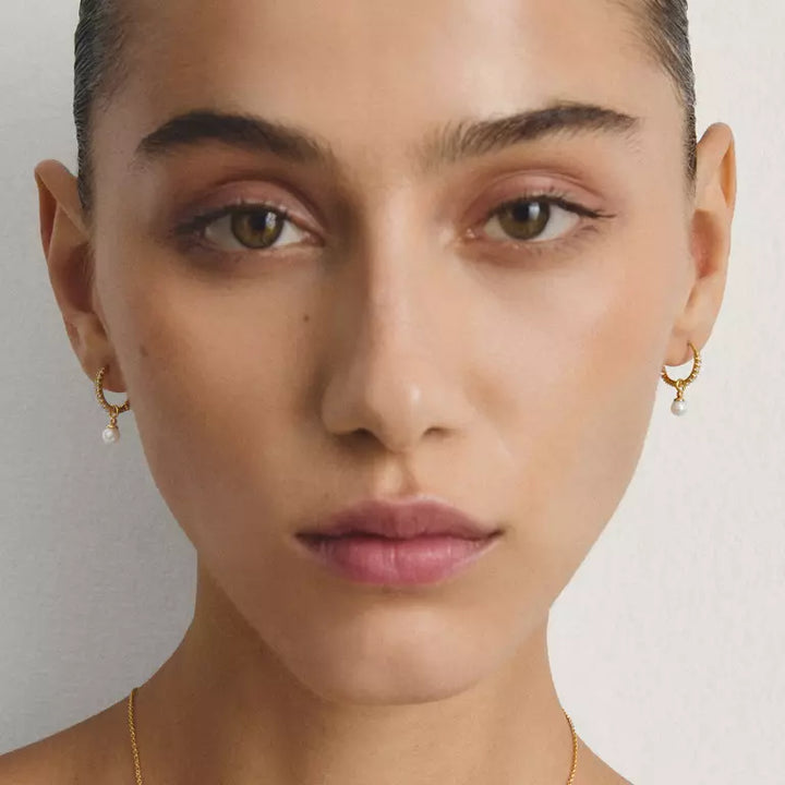 Close-up portrait of a model wearing small gold hoop earrings with dangling pearl-like beads, against a plain light background.