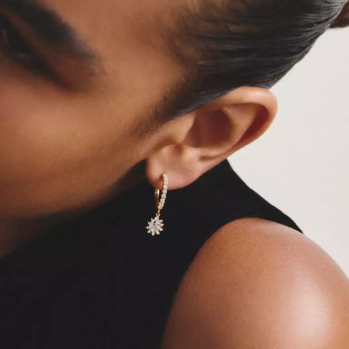A close-up of a person wearing a delicate gold huggie hoop earring adorned with small clear pavé stones and a dangling gold flower-like charm, paired with a black sleeveless top against a light neutral background.