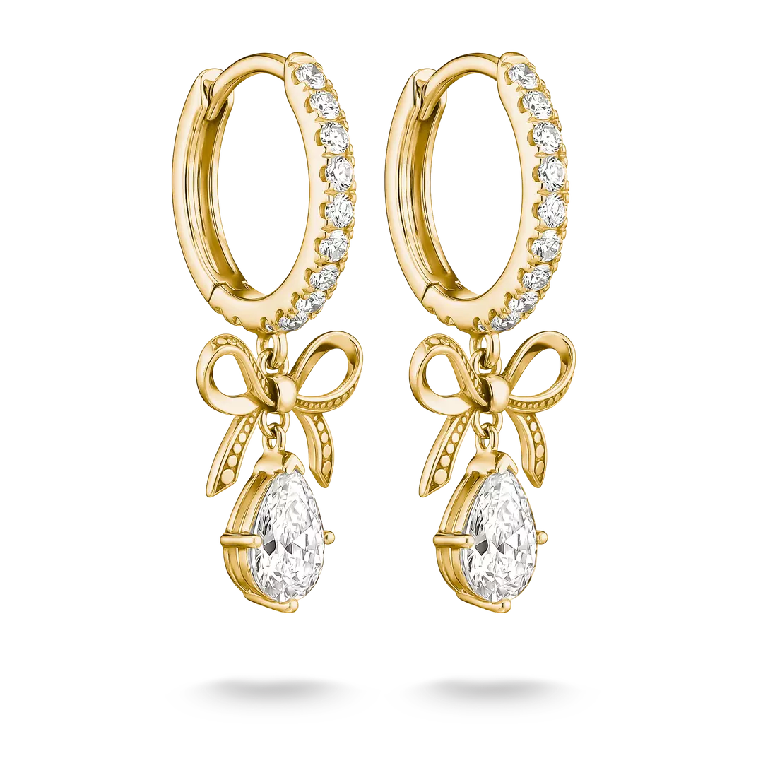 A pair of gold huggie hoop earrings with clear pavé stones along the top band, dangling bow motifs, and teardrop-shaped clear gemstones at the bottom, shown on a white background.