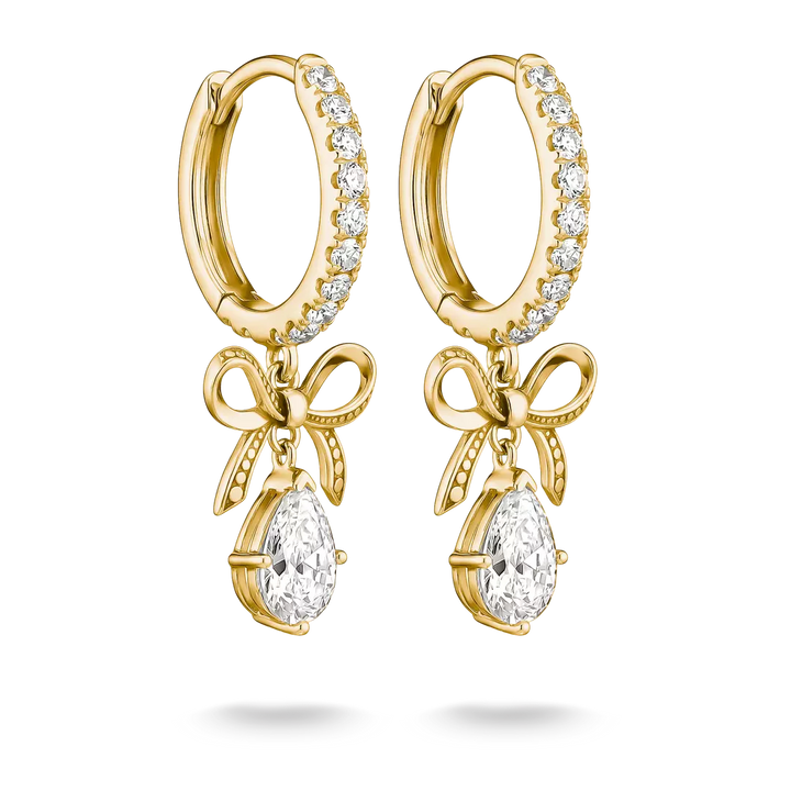 A pair of gold huggie hoop earrings with clear pavé stones along the top band, dangling bow motifs, and teardrop-shaped clear gemstones at the bottom, shown on a white background.
