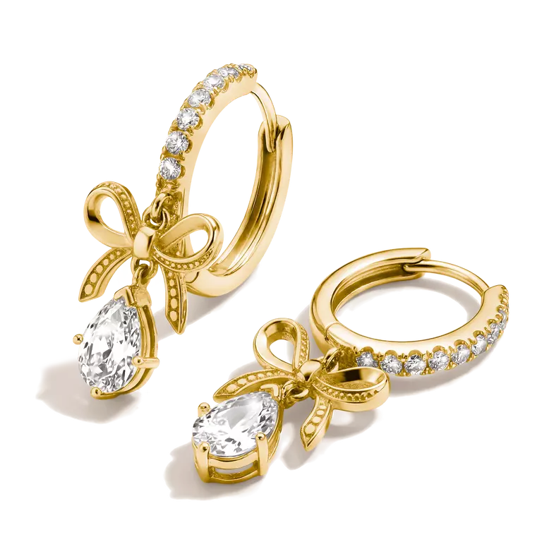 A pair of gold huggie hoop earrings with a bow and teardrop crystal charm, set against a clean white background.