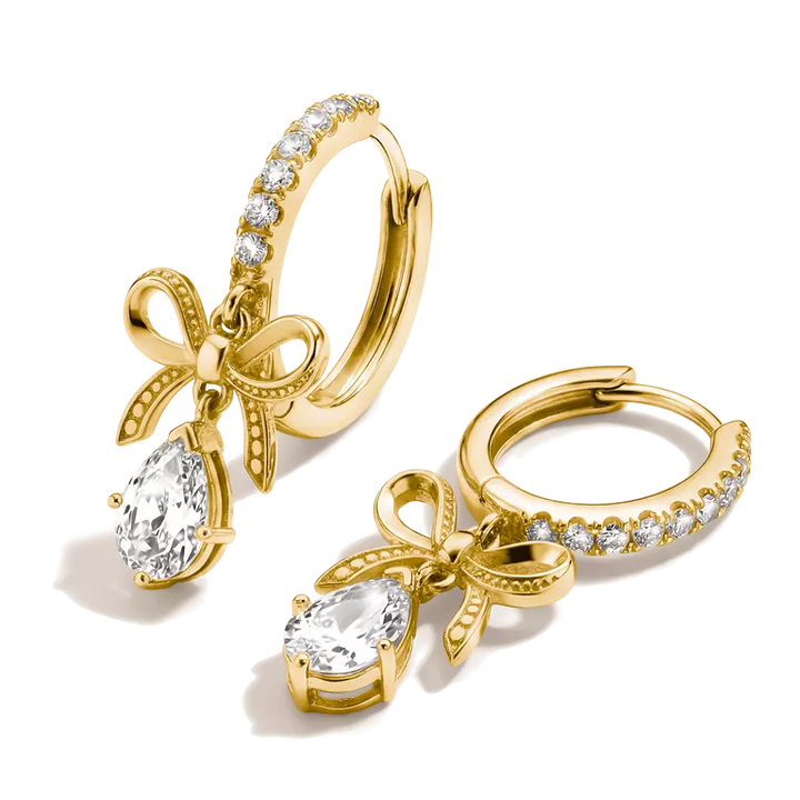 A pair of gold huggie hoop earrings with a bow and teardrop crystal charm, set against a clean white background.