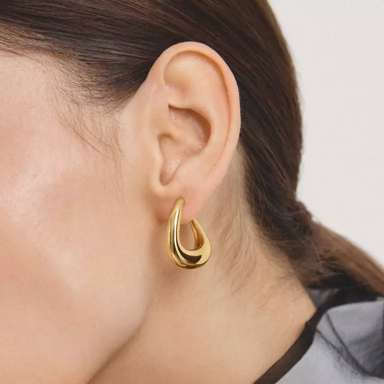 A close-up of a model's ear wearing a polished gold crescent-shaped hoop earring.