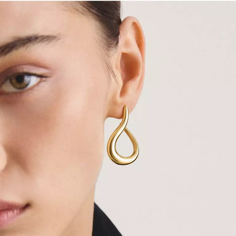 A close-up of a model's ear wearing a polished gold teardrop-shaped hoop earring against a light neutral background.