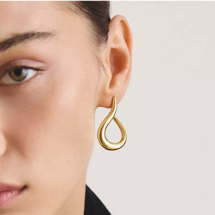 A close-up of a model's ear wearing a polished gold teardrop-shaped hoop earring against a light neutral background.