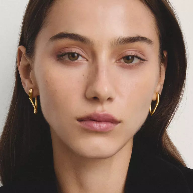 A close-up portrait of a woman wearing small gold hoop earrings against a plain light background.