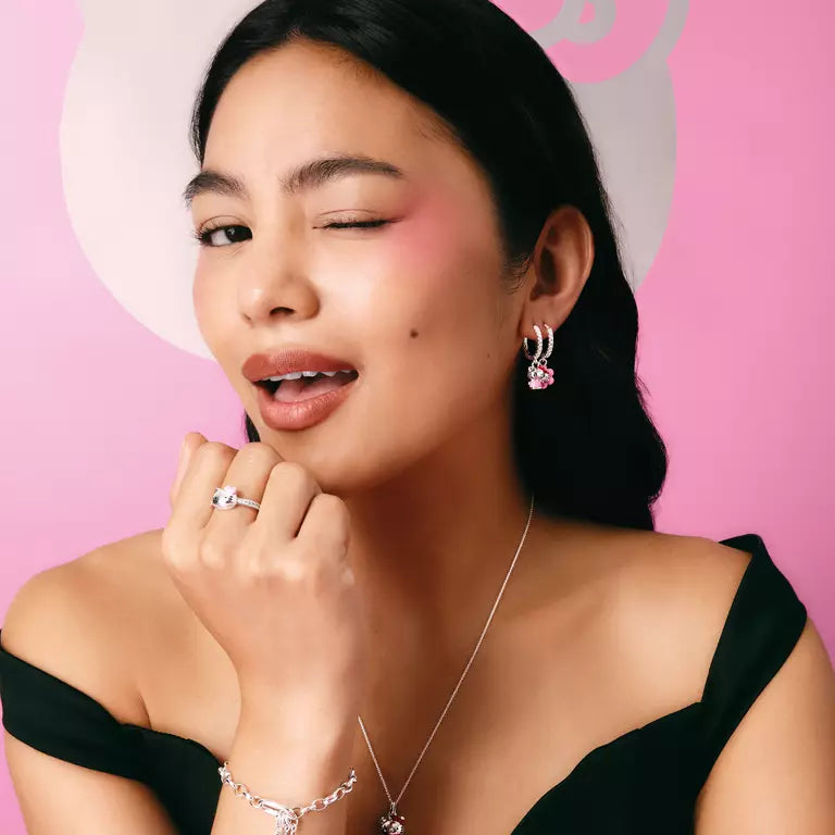 A close-up portrait of a woman with dark hair against a solid pink background, wearing a silver ring with a clear gemstone and a matching silver huggie hoop earring with pink flower charms.