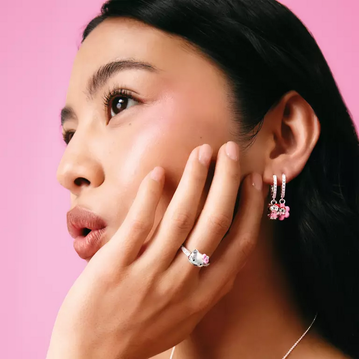 A close-up portrait of a woman with dark hair against a solid pink background, wearing a silver ring with a clear gemstone and a matching silver huggie hoop earring with pink flower charms.
