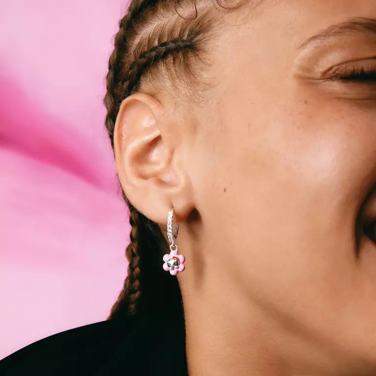 A close-up of a person wearing a small silver huggie hoop earring with a dangling pink flower charm, set against a solid pink background.