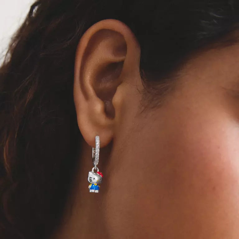 A close-up of a person's ear wearing a silver huggie hoop earring with a dangling Hello Kitty character charm.