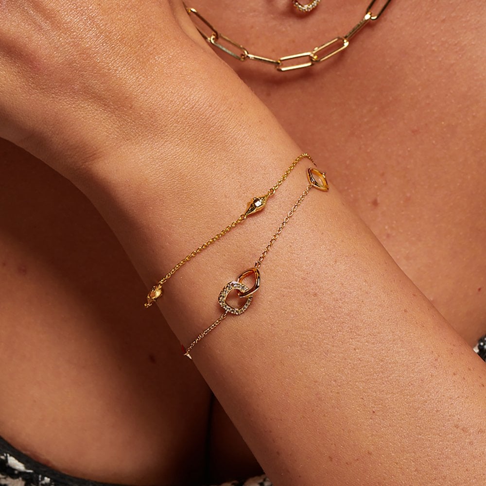 Gold bracelet on a wrist with a close-up of skin and additional jewelry in the background.