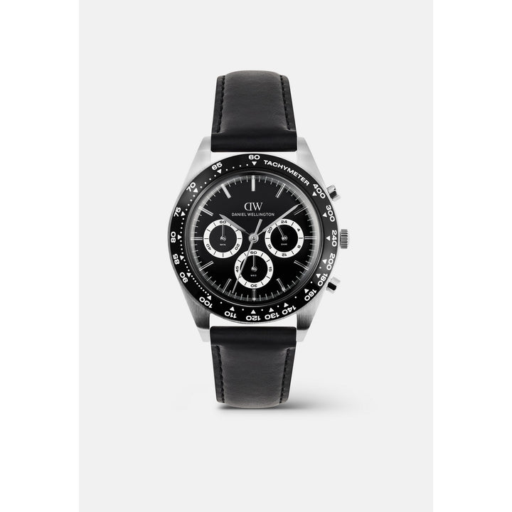 A wristwatch with a black dial, black leather strap, and a silver-toned stainless steel case. The dial features three subdials, a black tachymeter bezel, and the 'DW' logo for Daniel Wellington.