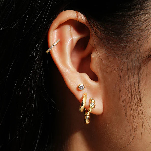 Close-up of an ear wearing gold hoop earrings with small stones on a dark background
