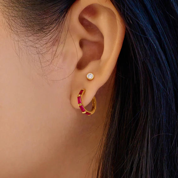 Close-up of an ear wearing gold hoop earrings with red gemstones and a diamond.