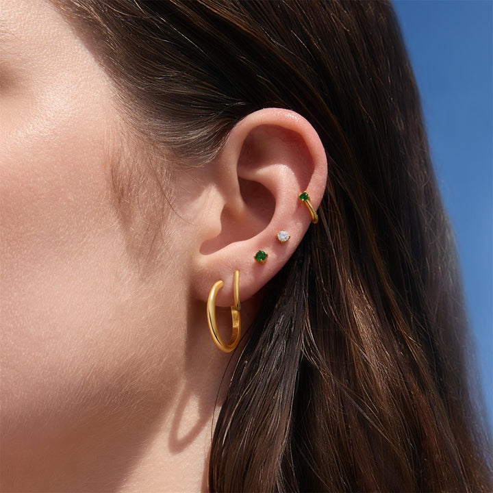 Close-up of an ear wearing gold hoop earrings with green gemstones against a blue sky.