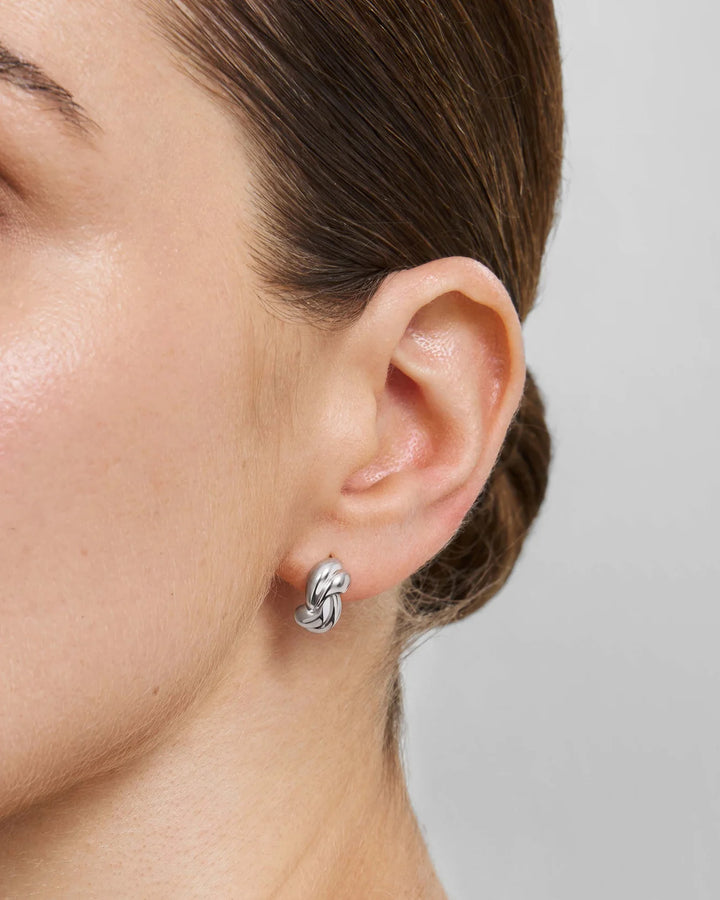 A close-up of a model's ear wearing a small silver knot stud earring against a light gray background.