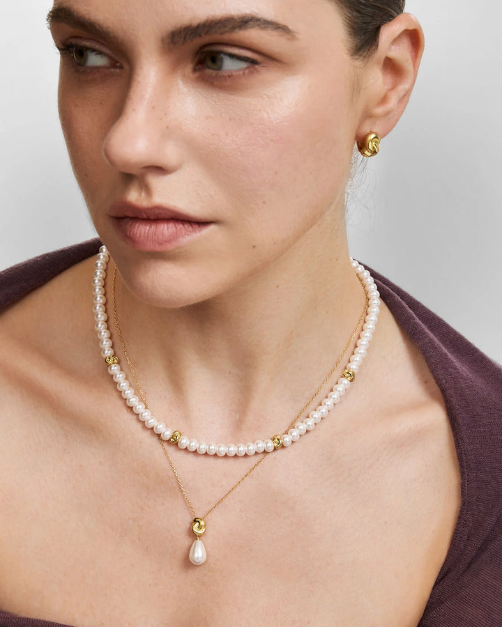 A close-up of a model wearing a layered necklace set featuring a short pearl strand with gold spacer beads and a delicate gold chain with a teardrop pearl pendant.
