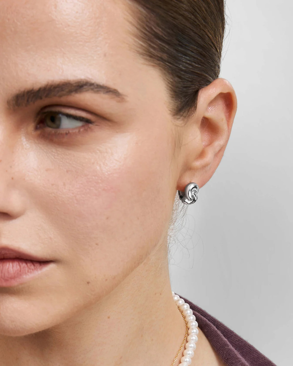 Close-up of a model's ear wearing a small silver spiral stud earring, with her face, neck, and a pearl necklace visible against a light gray background.