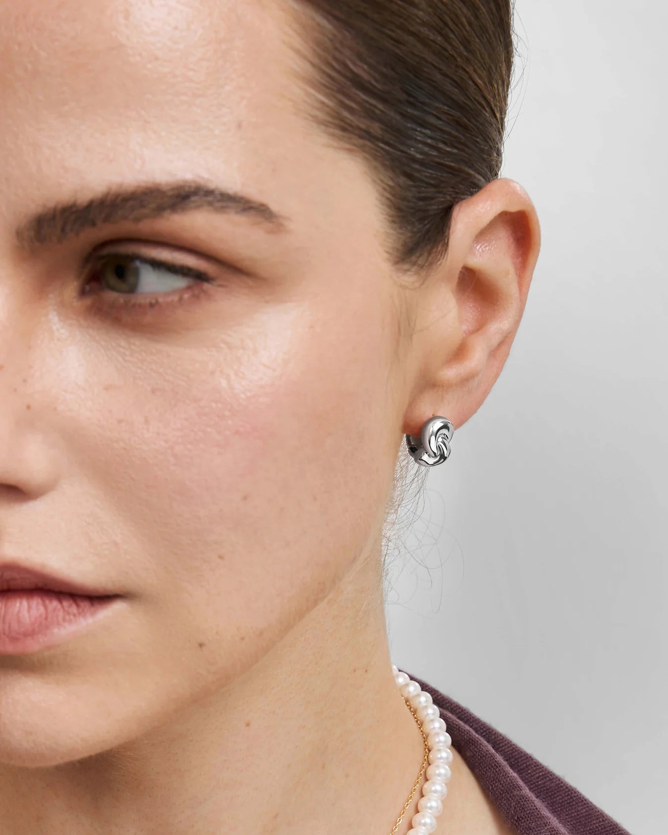 Close-up of a model's ear wearing a small silver spiral stud earring, with her face, neck, and a pearl necklace visible against a light gray background.